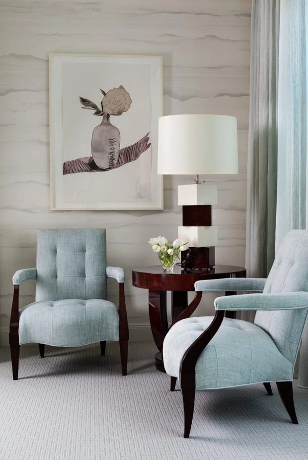A refined sitting area with two pale blue upholstered lounge chairs, dark wood frames, a round dark wood side table, and a sculptural table lamp. The room features a softly veined light wall finish, sheer blue-grey curtains, a pale textured carpet, and framed botanical artwork. The overall aesthetic is elegant, calm, and softly traditional with Art Deco-inspired furniture forms.