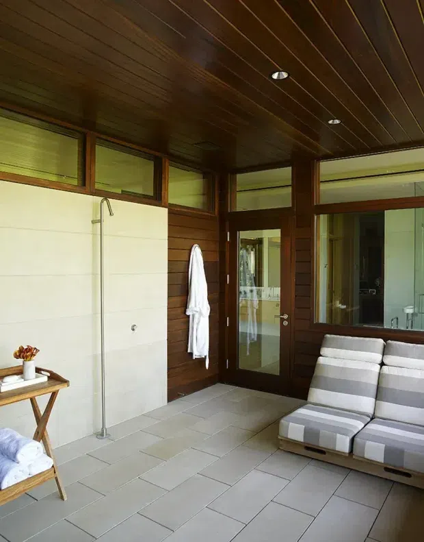 A warm, enclosed indoor-outdoor bathing and lounging space with dark timber wall and ceiling panelling, large glazed doors and windows, a tiled floor, an open shower area, and striped reclining lounge chairs. The room has strong natural light and a relaxed resort-like character.