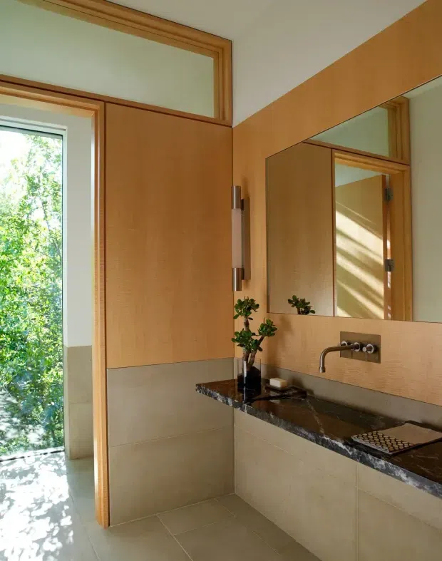 A compact, warmly lit bathroom with a wall-mounted dark stone vanity benchtop, integrated basin and brushed metal wall-mounted tapware. Large rectangular mirrors, honey-toned timber cabinetry and door frames, beige tiled walls and floor, and a narrow window overlooking greenery define the space. The room has clean-lined contemporary detailing with abundant natural light.