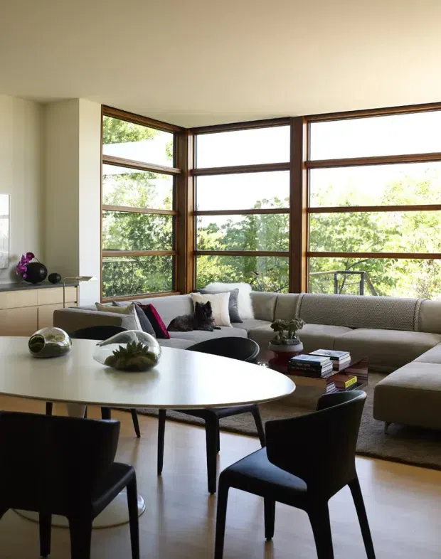 A bright, contemporary open-plan living and dining area with a large cream sectional sofa arranged around a low coffee table, alongside a rounded white dining table with black chairs. Floor-to-ceiling timber-framed windows wrap around the corner, bringing in abundant natural light and views of greenery. The room includes understated decorative objects, books, cushions, and indoor plants, creating a refined yet comfortable atmosphere.