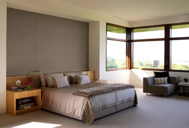 A spacious, softly lit bedroom with a large upholstered bed, integrated timber bedside and storage joinery, a full-height grey feature wall, and expansive dark timber-framed corner windows overlooking greenery. A lounge chair and side table sit beside the windows, creating a calm, refined retreat.