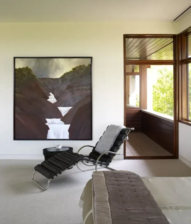 A calm, minimalist bedroom with pale walls and carpet, featuring a black leather chaise lounge, a large framed landscape artwork, and a bed in the foreground. Timber-framed glazing and a partially enclosed sunroom or balcony-like space open to leafy surroundings.