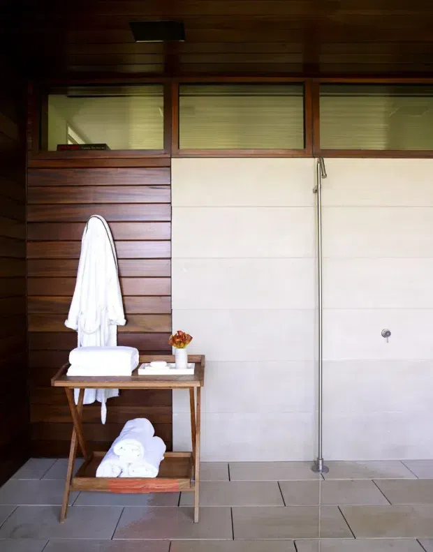 A compact, dark-toned bathroom with a walk-in shower occupying the right side. The room features large pale wall tiles, matching floor tiles, horizontal dark timber wall panelling, upper timber-framed clerestory windows with blinds, and a simple wooden towel table holding folded white towels and toiletries. The atmosphere is warm and restrained, with natural timber contrasting against the light tile surfaces.