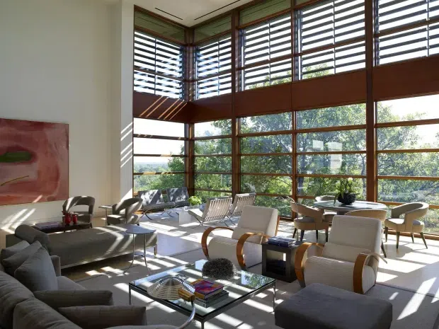 A bright, expansive double-height living room with extensive timber-framed glazing and horizontal louvres. The space contains a large sectional sofa, sculptural lounge chairs, glass coffee tables, and a dining area at the rear. Strong sunlight creates dramatic linear shadows across the pale carpet and furnishings, while the surrounding greenery is visible through the windows.