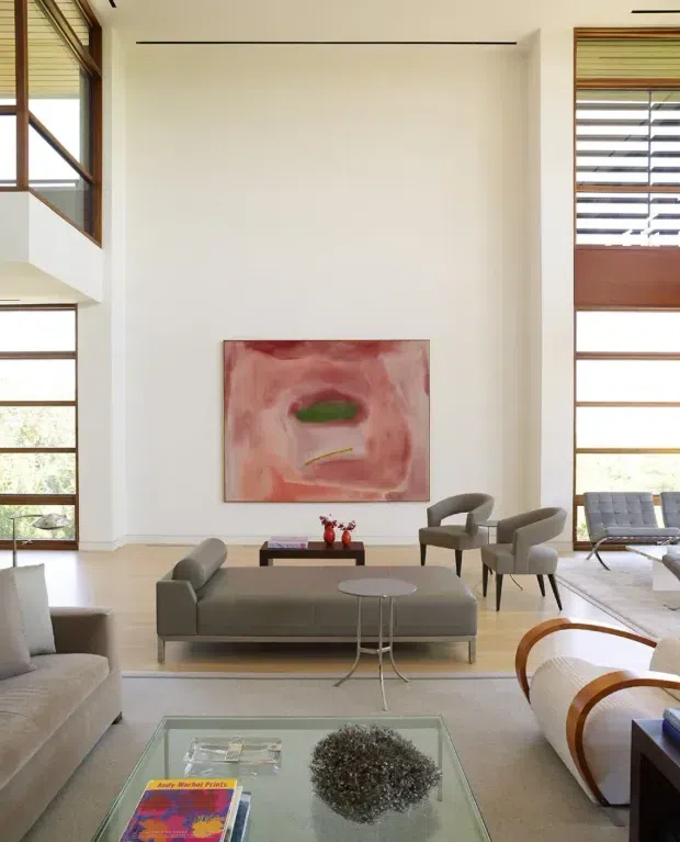 Bright, spacious contemporary living room with very high ceilings, expansive timber-framed windows, horizontal timber louvres, pale walls and light flooring. A large grey sofa, armchairs, glass coffee table, and sculptural lounge chairs surround a low central table. Abstract artwork adds a muted pink and green focal point, while abundant glazing creates an airy, gallery-like atmosphere.
