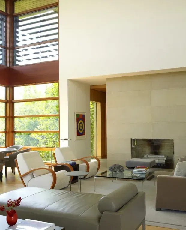 A bright, double-height living room with expansive timber-framed glazing and horizontal louvres overlooking greenery. The room features a pale stone or plaster fireplace wall, contemporary upholstered seating, a large light-toned sectional sofa, glass coffee table, and neutral finishes. The high ceiling and abundant natural light create an airy, refined atmosphere.