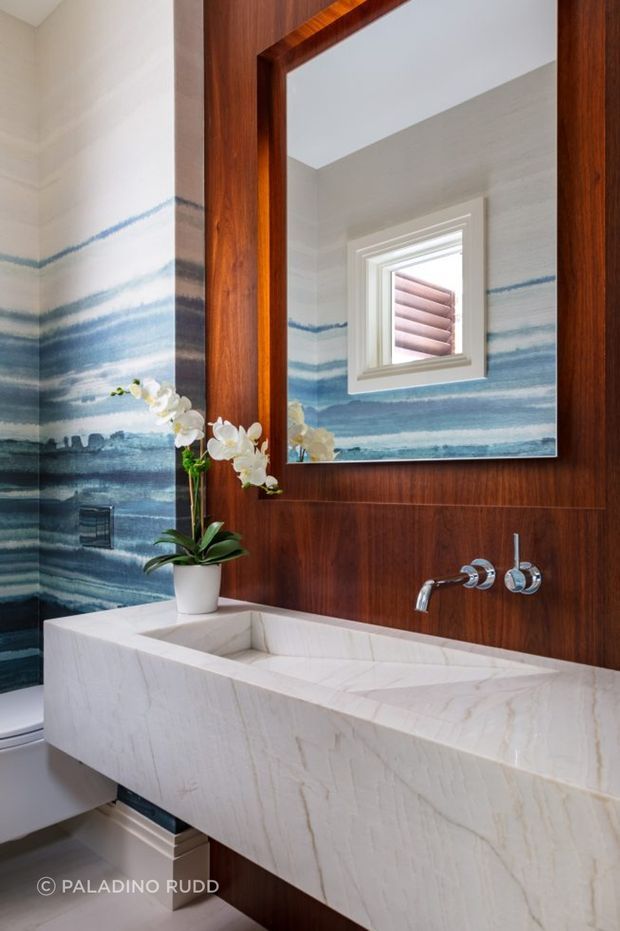 A refined bathroom featuring a sculptural white stone trough basin with wall-mounted chrome tapware, set against rich dark timber wall panelling. The walls are finished with a dramatic blue and white wave-patterned surface, echoed in the large mirror. A potted white orchid adds a natural decorative touch, while a small high window provides daylight. The space has a luxurious, coastal-inspired feel with strong material contrast between stone, timber, chrome and blue patterned walls.