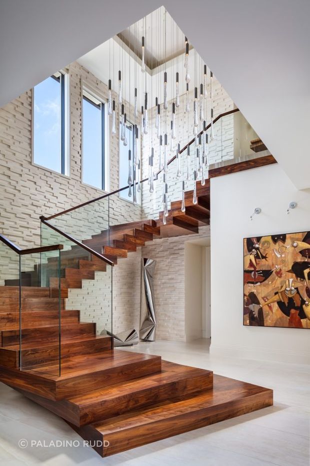 A bright, dramatic modern stairway and landing with a sculptural floating staircase in rich dark timber, clear glass balustrades, and slender dark handrails. The stair rises through a double-height void lined with textured white brick walls and large vertical windows. Numerous suspended pendant lights hang above the stair, while a timber mezzanine and contemporary artwork add visual interest. Pale flooring and white walls create a high-contrast, gallery-like interior.