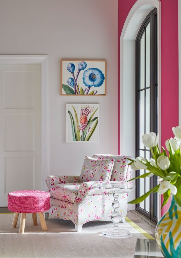 A bright, colourful sitting area featuring a floral upholstered armchair beside a tall arched black-framed window, a pink textured stool, framed botanical artwork, and a large vase of white tulips. The room has pale walls, white panelled doors and trim, and a patterned floral furnishing scheme with a cheerful, eclectic feel.
