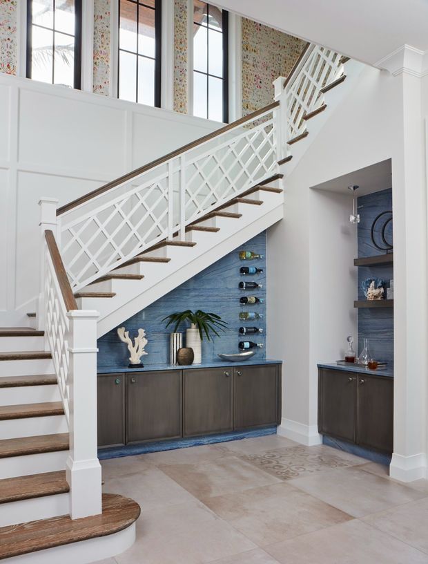 Bright, refined stairway and foyer featuring a white painted staircase with distinctive diamond-pattern balustrades and warm timber stair treads and handrail. Tall black-framed windows and floral wallpaper rise above white wall panelling. Beneath the stairs is a built-in dark timber bar and storage area with a blue textured stone or tile feature wall, wine bottle storage, shelving, and decorative objects. The floor is finished in large pale stone-look tiles.