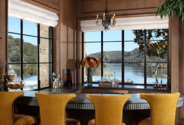 A richly panelled dining or entertaining space with a dark stone-topped built-in counter beneath large black-framed windows overlooking a calm waterfront landscape. Four mustard-yellow upholstered bar chairs line the counter, while a decorative chandelier, floral arrangement, glassware, and indoor plants add refined character. Warm timber joinery and trim frame the windows, creating an elegant, traditional interior with strong natural light and water views.
