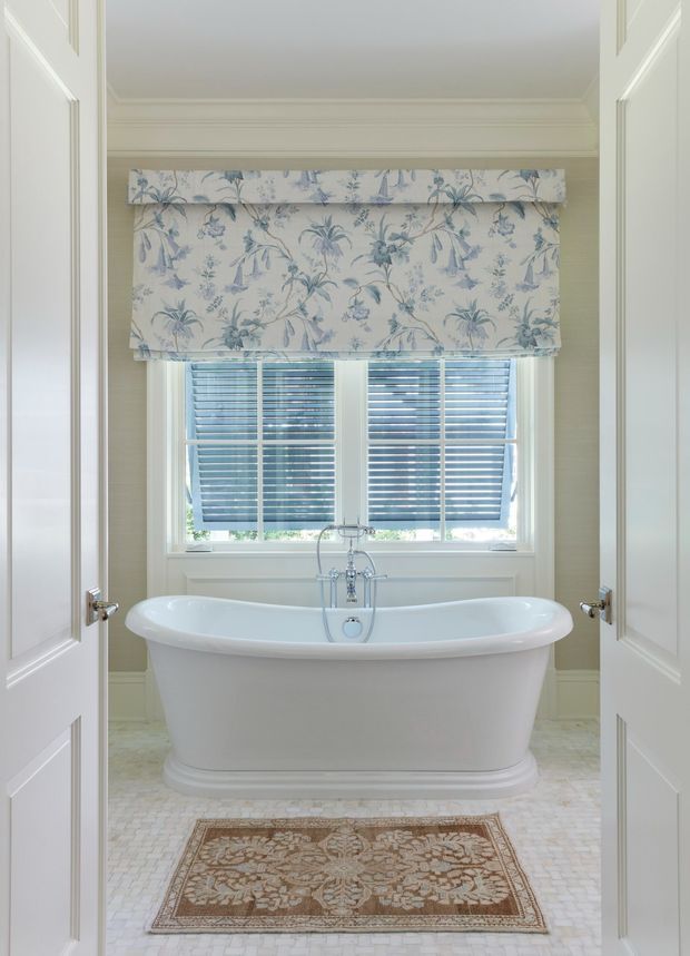 Bright, refined bathroom viewed through open double doors, featuring a freestanding white clawfoot-style bathtub beneath a large window with plantation shutters and a blue floral Roman blind. The room has pale neutral walls with traditional moulding, a small mosaic tile floor, and a patterned area rug.