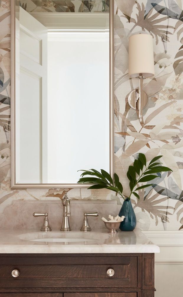 A refined bathroom vanity vignette featuring a dark timber vanity with drawers, a pale stone countertop and integrated basin, polished metal three-piece tapware, a large framed mirror, botanical patterned wallpaper, white wall panelling, and a chrome wall sconce. A teal vase with leafy greenery adds contrast.