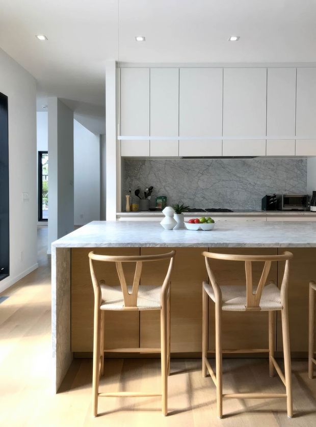 A bright, contemporary kitchen centred on a large pale stone island with integrated seating. The kitchen features flat-panel white overhead cabinetry, warm light-wood island cabinetry, and a dramatic grey-veined stone splashback. Two light timber bar stools sit at the island, with recessed ceiling lights and a hallway or living area visible beyond.