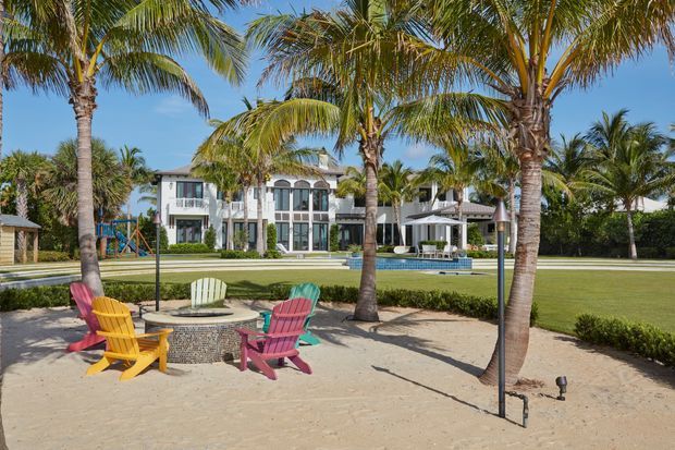 A tropical beachfront-style outdoor courtyard or resort landscape with sandy seating areas, palm trees, colourful Adirondack chairs, manicured lawns, and a large white multi-storey residence in the background. A swimming pool and covered outdoor entertaining area are visible beyond the lawn.