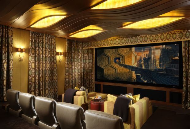 A richly decorated home cinema with rows of plush upholstered theatre seating facing a large wall-mounted screen. The room features dark timber panelling, patterned geometric wall coverings, warm amber integrated ceiling lighting, and an ornate, sculptural curved ceiling with illuminated wave-like panels. Additional lounge seating and small tables sit behind the main theatre chairs, creating a luxurious, intimate entertainment space.