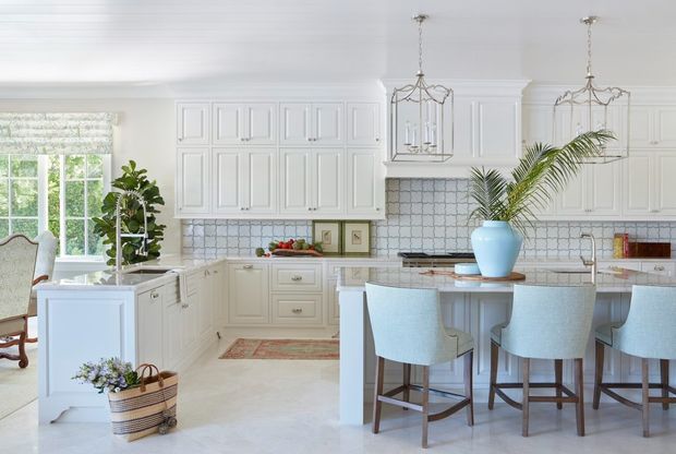 Bright, spacious traditional kitchen with extensive white raised-panel cabinetry, a large central island with seating, a pale blue tiled backsplash, marble-look benchtops, and decorative hanging lanterns. A dining area and indoor plants are visible to the left.