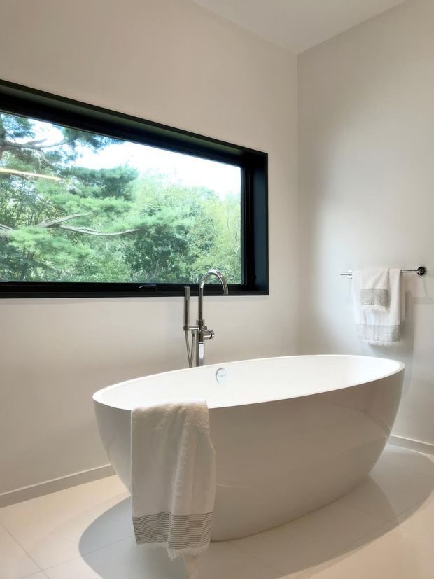 Bright, minimalist bathroom featuring a freestanding white oval bathtub beneath a large black-framed window overlooking trees. A chrome floor-mounted tub filler and simple wall-mounted towel rail with white towels complete the calm, neutral space. Pale tiled flooring and off-white walls create an airy, contemporary setting.