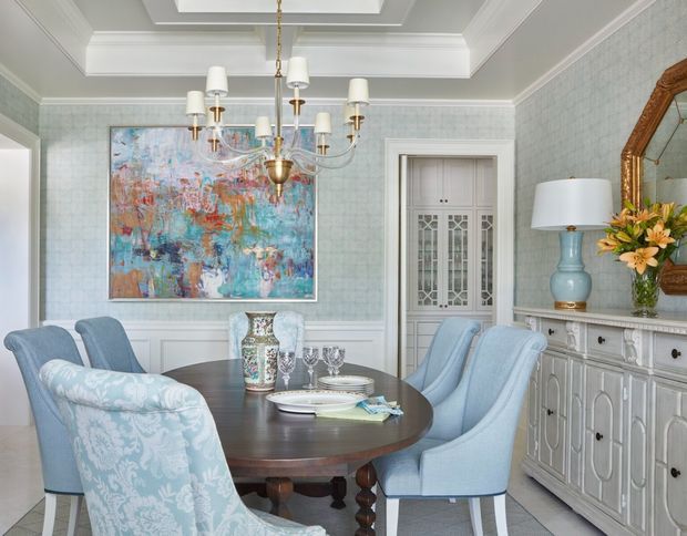 Elegant dining room with a round dark wood dining table surrounded by upholstered blue dining chairs, pale blue patterned wallpaper, white wainscoting and detailed coffered ceiling. A central brass-and-glass chandelier, ornate sideboard, decorative mirror, floral arrangement and colourful abstract artwork add refined traditional character.