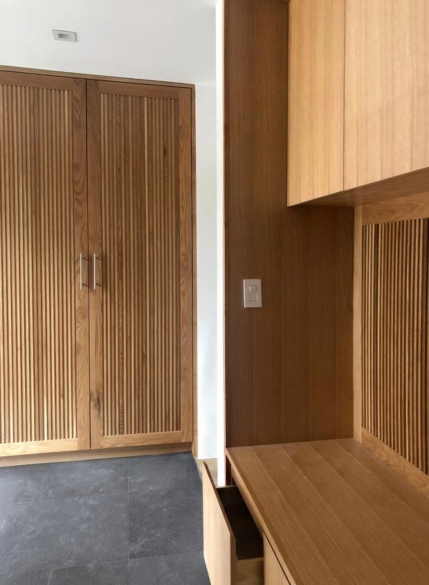 A close-up interior view of a custom timber joinery area, with tall fluted wood-panelled cupboard doors on the left and matching timber cabinetry, wall panelling, and a built-in bench or desk surface on the right. The space has a white ceiling and wall sections, dark large-format floor tiles, and minimal brushed metal hardware. The strong vertical fluting and warm wood tones create a refined, contemporary built-in feature.