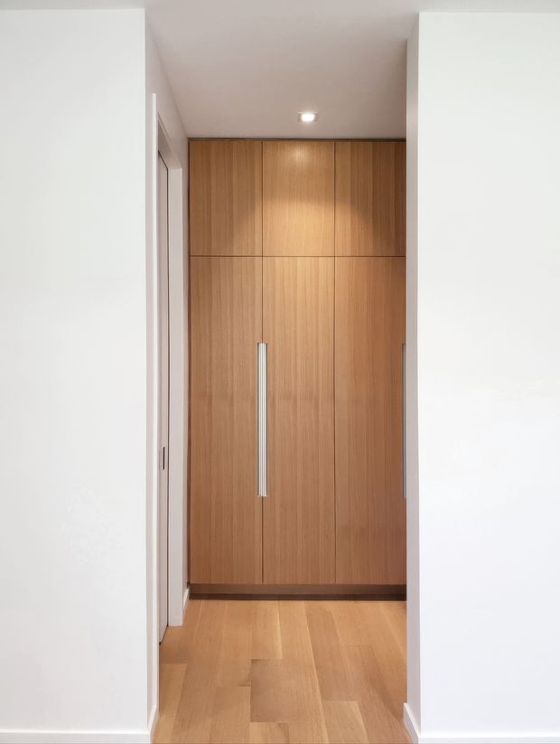 A narrow, clean-lined hallway or entry passage viewed through a doorway, featuring full-height built-in timber cabinetry with tall vertical silver handles at the far end. The space has white walls, a recessed ceiling light, and warm timber flooring. The cabinetry and flooring create a restrained, contemporary interior with a warm neutral palette.