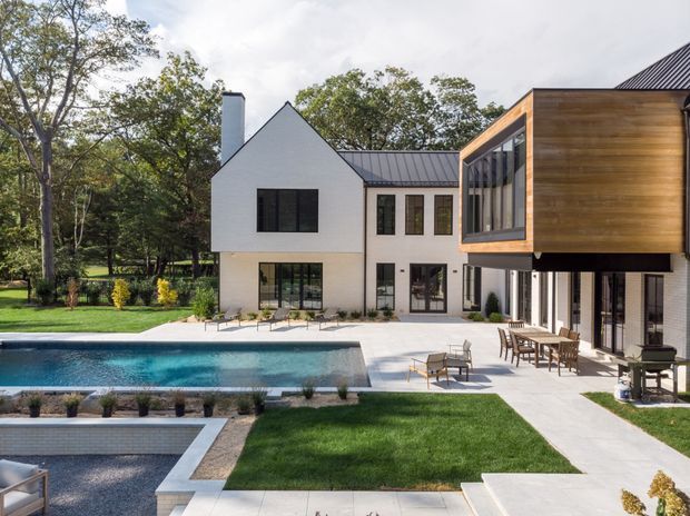 A contemporary multi-wing residential home surrounding a landscaped backyard and swimming pool. The white gabled main volume contrasts with a projecting timber-clad upper-level box and dark metal pitched roofs. Large glazed openings connect the interior to a pale paved terrace with outdoor dining and lounge furniture, while mature trees and manicured lawns frame the property.