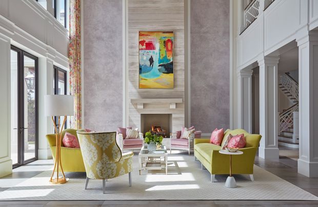 Bright, sophisticated double-height living room with a large fireplace feature wall, tall glazed doors, gallery artwork, built-in upper-level balustrades, and a visible staircase. The room is furnished with lime-green and blush-pink seating, a patterned accent chair, glass coffee table, and a large pale rug. The overall scheme is colourful, refined, and contemporary with classical architectural detailing.
