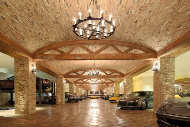 A grand interior car display and storage space with rows of luxury vehicles arranged beneath a vaulted brick ceiling. Exposed timber trusses span between substantial stone-clad columns, while repeated wrought-iron chandeliers and wall sconces provide warm illumination. The floor is finished in terracotta-toned patterned tile, and large mirrors reflect the vehicles and architectural structure.