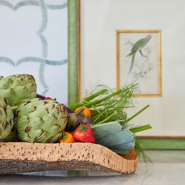 A close-up still life of artichokes, tomatoes, citrus and leafy vegetables arranged on a thick natural bark-edged wooden serving board. The arrangement sits on a pale countertop, with a green-trimmed framed botanical artwork and a framed bird illustration in the background.