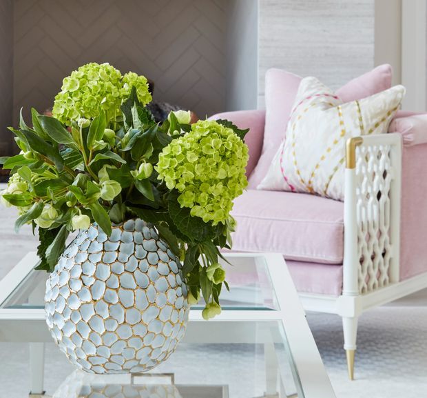 A close-up interior vignette featuring a large pale blue and gold decorative vase filled with green hydrangeas on a glass-topped coffee table. Behind it is a pale pink upholstered armchair with patterned cushions, white lattice-style arms, and gold-toned tapered feet. The room has a refined, softly coloured decorative aesthetic with a dark chevron-patterned wall and light textured wall finish.