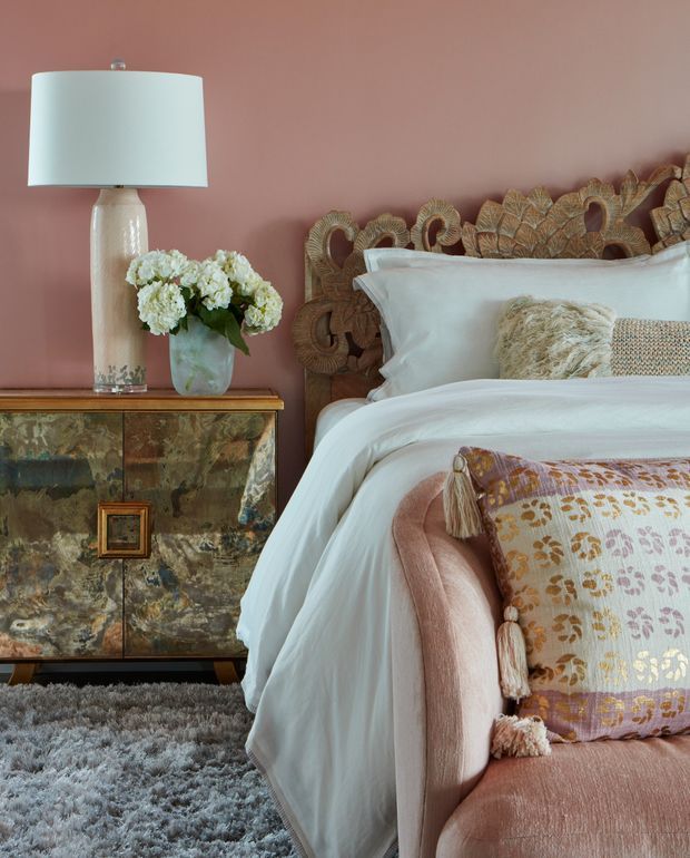 A styled bedroom vignette with a carved natural-wood headboard, white bedding, blush pink upholstered bed, patterned decorative cushions, and a warm dusty-rose wall. A marble-patterned timber sideboard with brass-toned detailing supports a ceramic lamp and a vase of white flowers. A soft grey rug covers the floor.