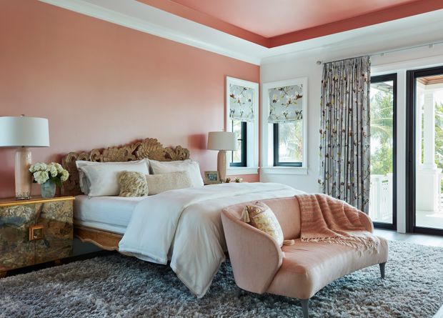 A luxurious bedroom with a blush-pink accent wall, ornate upholstered bed, white bedding, and a curved blush lounge chair. Patterned curtains frame tall black-framed glazed doors and windows, while a textured grey rug, mirrored bedside cabinetry, lamps, and layered cushions complete the refined interior.