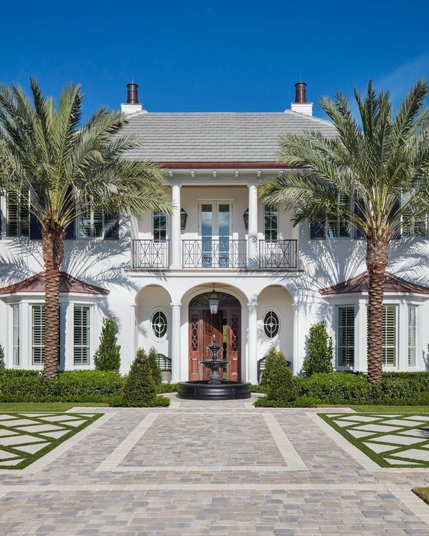 Formal front elevation of a grand two-storey white stucco residence with a symmetrical composition, grey hipped roof, twin chimneys, shuttered windows, columned balcony, arched entry portico, dark timber front doors, and a central fountain. Palm trees, clipped hedges, geometric lawn panels, and a broad patterned stone driveway create a resort-like landscaped approach.
