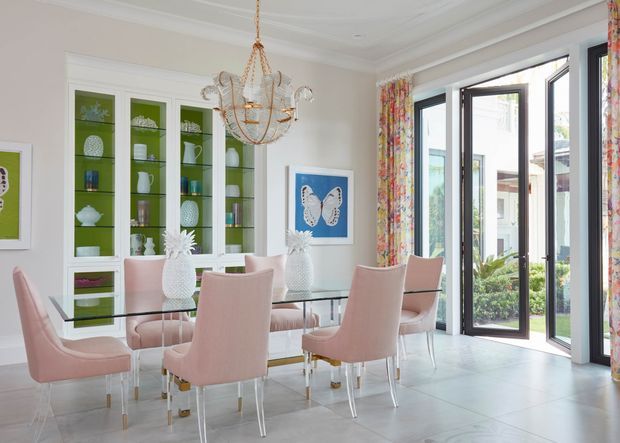 Bright, elegant dining room with a long glass dining table surrounded by blush-pink upholstered chairs. A large white built-in display cabinet with green backing occupies one wall, while full-height glazed folding or bi-fold doors open to an outdoor area. The room features white walls and ceiling mouldings, pale tiled flooring, colourful floral curtains, butterfly artwork, and a decorative crystal chandelier.