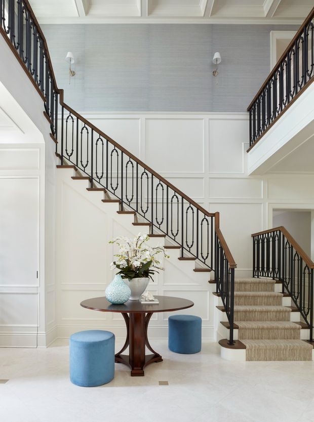 Elegant residential stair hall with a sweeping bifurcated staircase, dark wrought-iron balustrades and warm timber handrails. The walls feature white raised-panel wainscoting and pale textured wallcovering, while the ceiling has decorative coffered beams. A small dark wood pedestal table with blue upholstered stools and floral decor sits beneath the stairs, creating a refined, traditional transitional entry space.