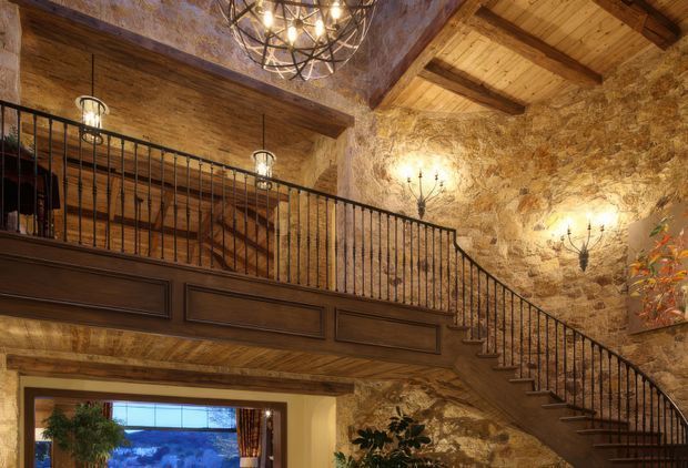 Rustic, warmly lit interior featuring a high vaulted timber ceiling with exposed beams, rough natural stone walls, a dark timber mezzanine or landing, and a sweeping staircase with black metal balustrades. Decorative wrought-iron chandeliers and wall sconces reinforce the historic lodge or European farmhouse character. A lower living area is partially visible beneath the mezzanine.