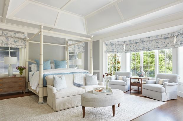 Bright, elegant bedroom with a four-poster bed, upholstered bench, armchairs, and a round ottoman arranged in a spacious sitting area. The room features pale grey walls, white architectural trim, a coffered and paneled ceiling, large multi-pane windows, patterned Roman shades, light carpeting, and dark timber flooring at the perimeter. The overall styling is refined and traditional with a soft blue, white, and grey palette.