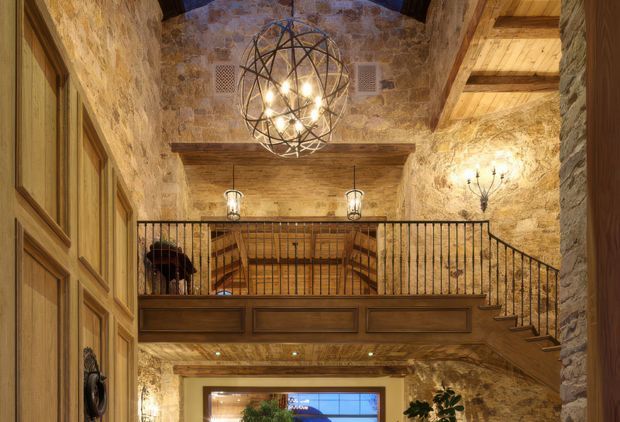 A rustic, double-height interior entry or foyer with exposed rough stone walls, heavy timber beams and ceiling boards, paneled timber doors, and a timber mezzanine reached by a staircase. Black wrought-iron balustrades and dramatic spherical and wall-mounted lighting add a refined lodge-like character.