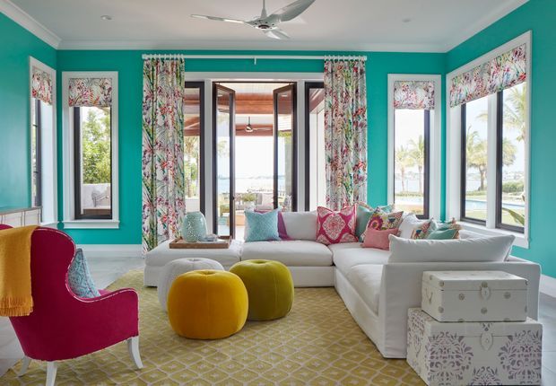 A bright, colourful living room with vivid turquoise walls, large black-framed windows and glazed doors, patterned floral curtains and a white sectional sofa. Jewel-toned cushions, mustard upholstered ottomans, a magenta accent chair, and a patterned area rug create a playful, eclectic interior. The central doors open to an outdoor view with a pool or water feature beyond.