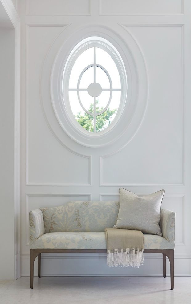 Bright, refined interior vignette featuring a pale upholstered two-seat settee with patterned fabric, a neutral throw, and a cushion. The settee sits against a white wall with detailed raised panel moulding and a distinctive oval window with radial muntins. The space has a calm, elegant, classic character with light flooring and abundant daylight.