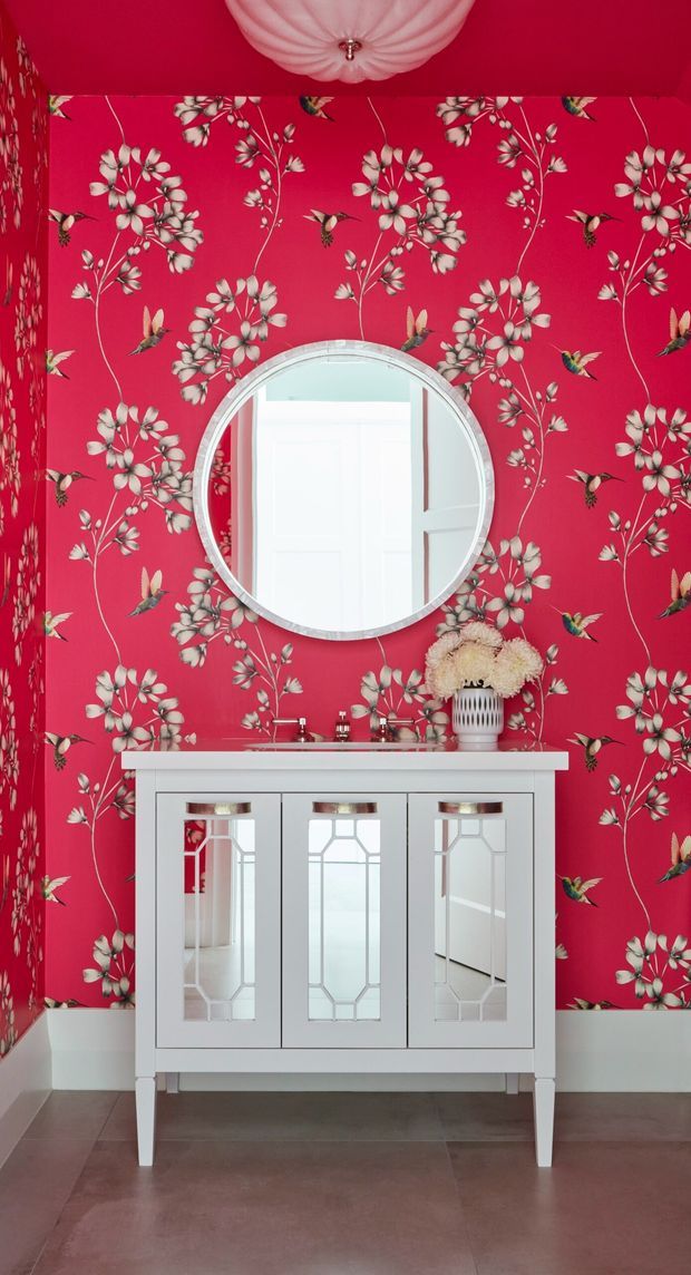 A compact, highly decorative bathroom or powder room with vivid fuchsia-red floral and hummingbird wallpaper covering the walls and ceiling. A white mirrored vanity with three inset mirrored cabinet doors sits beneath a large round wall mirror, with a small white basin, chrome tapware, and floral arrangement. The room has a white ceiling light, white trim, and a dark muted floor.