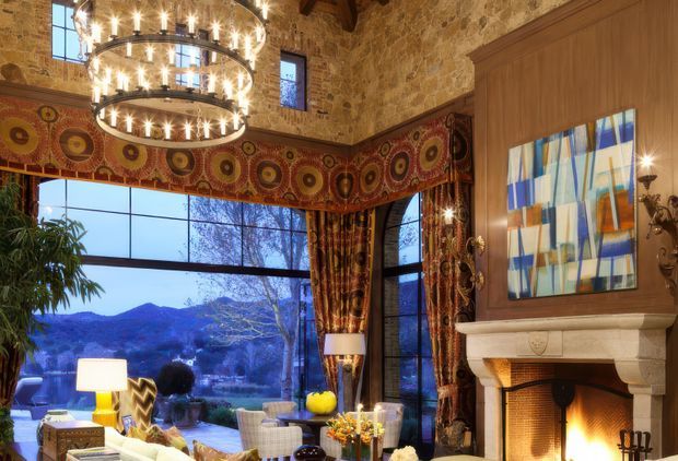 An ornate, double-height living room with exposed rustic stone walls, a large circular multi-tier chandelier, richly patterned geometric curtains and valances, and expansive corner glazing overlooking a mountainous landscape. A substantial carved stone fireplace with a timber surround and abstract artwork forms a major focal point, while upholstered seating and a coffee table create a comfortable lounge area.