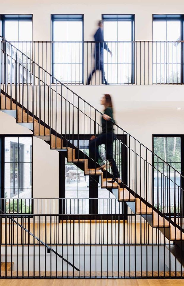 A bright multi-level interior atrium or circulation space with multiple flights of open-riser timber stairs and slim black metal balustrades. Large vertically aligned windows provide abundant natural light, while blurred figures move along the staircases and upper landing. The composition emphasises the strong geometric repetition of railings, stairs, and glazing.