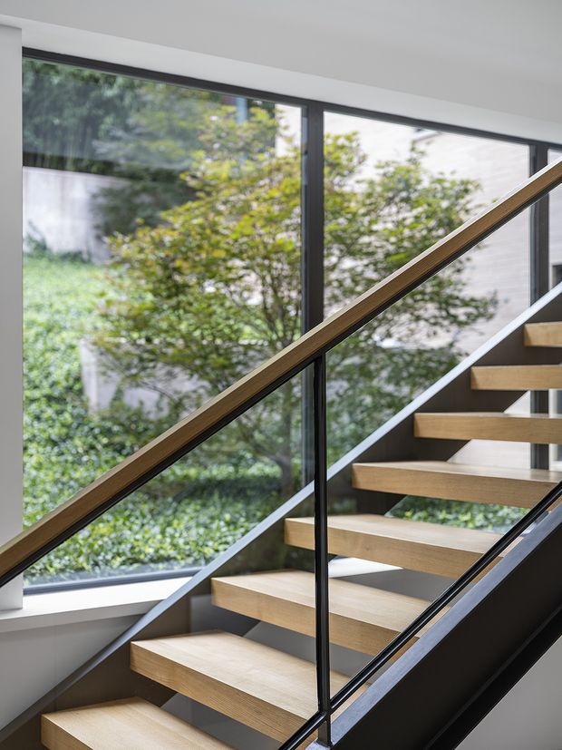 A contemporary interior stairway with open-riser timber treads, dark metal stringers, and slender black-framed glass balustrades. Large floor-to-ceiling windows beside the stairs provide abundant natural light and views of leafy greenery outside. The space has clean lines, neutral white and grey surfaces, and a refined minimalist appearance.