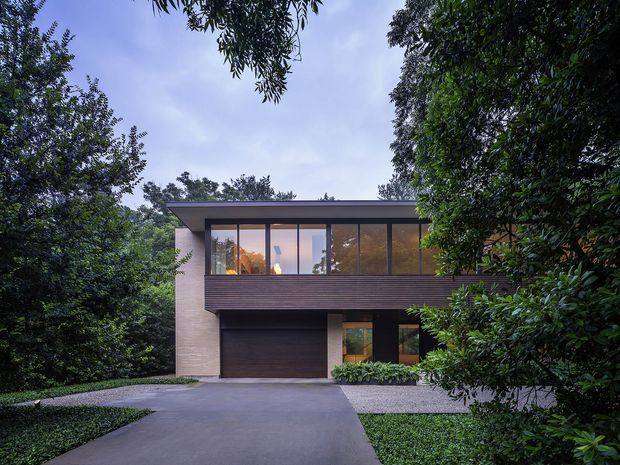 A contemporary two-storey residential house viewed from the driveway, nestled among mature trees and dense landscaping. The facade combines dark horizontal cladding, pale masonry, extensive upper-level glazing, and a broad low-pitched roof overhang. A garage and entry are visible at ground level, with a paved driveway leading toward the home.