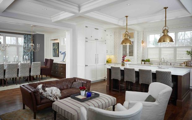A bright open-plan interior combining a traditional kitchen, dining area, and living space. The kitchen features white shaker-style cabinetry, a large island with seating, white subway-tile walls, dark wood flooring, and brass pendant lights. The adjacent dining and lounge areas include upholstered dining chairs, a leather sofa, armchairs, and an ottoman. Coffered ceilings, layered white trim, plantation shutters, and classic detailing create a refined, comfortable look.