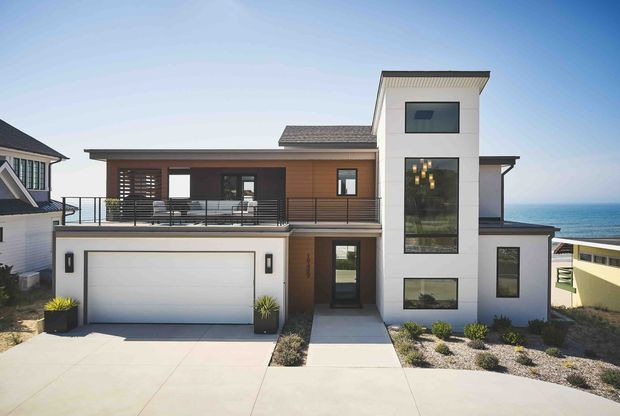Front exterior view of a contemporary multi-level coastal residence, featuring a flat-roofed white vertical tower with stacked dark-framed windows, warm timber-toned cladding, a broad upper balcony, and an integrated double garage. The ocean is visible beyond the property.