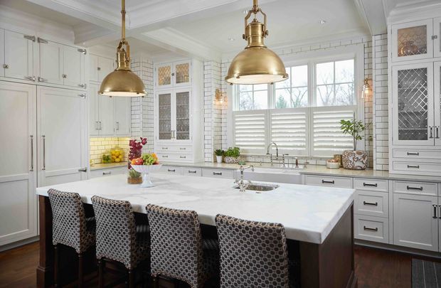 Bright, spacious kitchen featuring extensive white shaker-style cabinetry, white brick-pattern wall tiling, a large central island with integrated sink and seating for four, and polished brass pendant lights. Shuttered windows, glass-front display cabinets, dark timber flooring, and patterned upholstered bar stools create a refined traditional transitional interior.