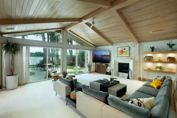 A spacious, bright living room with a vaulted timber-lined ceiling and exposed structural beams. Floor-to-ceiling glazing and sliding glass doors open to an outdoor terrace and landscaped setting. The room contains a large sectional sofa, additional lounge chairs, a central coffee table, a television, fireplace with light stone surround, and open display shelving. The overall design combines warm natural timber, pale finishes, and abundant daylight.
