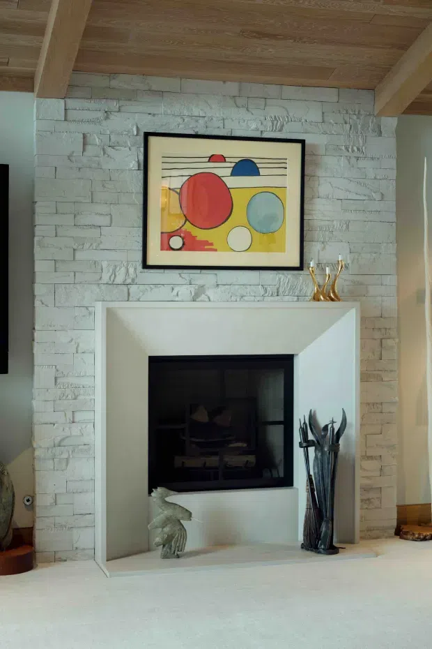 Interior view of a living room fireplace with a white-painted textured stone masonry surround and broad white mantel. The fireplace has a dark inset firebox, with decorative fireplace tools and sculptural objects arranged nearby. Above it hangs colourful geometric artwork in a black frame. The ceiling features warm timber lining and exposed timber beams, creating a rustic yet refined focal point.